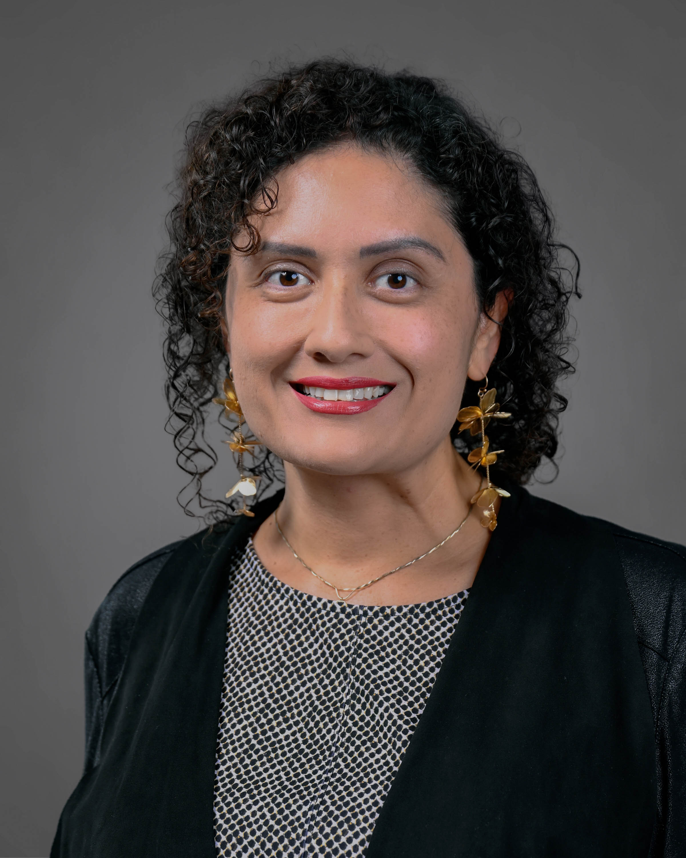 Kind looking woman smiling with curly dark hair in a dark jacket with a black and white shirt